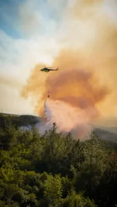 Green firefighting helicopter releasing a long water drop over a forested area with orange smoke from a wildfire behind it.