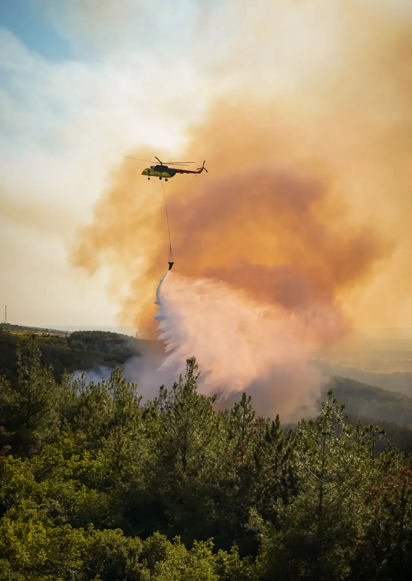 Green firefighting helicopter releasing a long water drop over a forested area with orange smoke from a wildfire behind it.